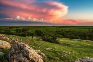 Expansive and serene view of the Flint Hills in Kansas, showing the characteristic undulating terrain covered in golden and green prairie grasses under a vibrant sky with scattered clouds and hues of blue and pink.