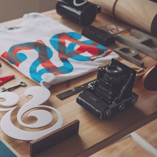 Close-up shot of a curated collection of design and vintage items, including an old folding camera, a graphic t-shirt, and a large white ampersand art piece, all arranged on a light wood desk.